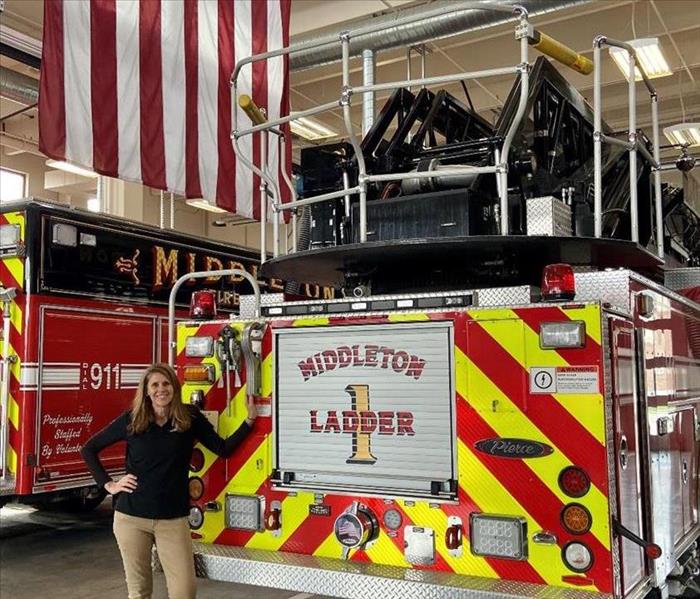 a woman in black SERVPRO shirt and khaki pants standing in front of fire trucks with American flag in background