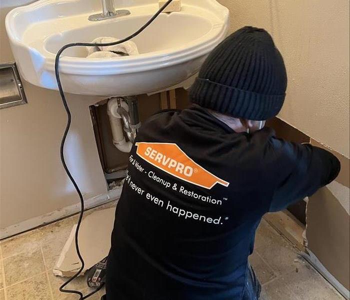 a man in a servpro uniform cutting drywall underneath a sink to access a broken water pipe 