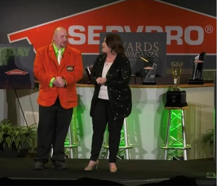 a man and a woman announcing SERVPRO Convention winners of accomplishment he is dressed in an orange sport coat 