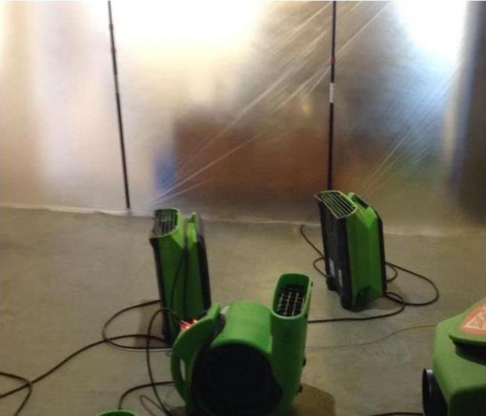 a concrete floor with three green air movers and a green dehumidifier contained within a plastic wall for containment 